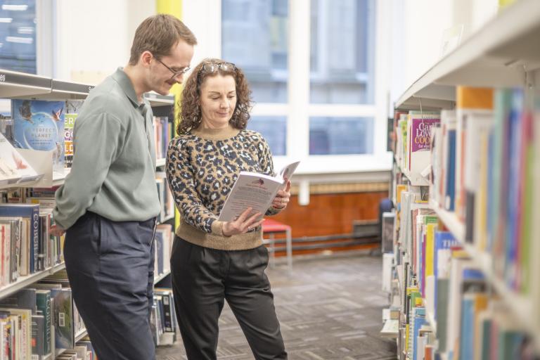 Two people looking at a book in between shelves of books