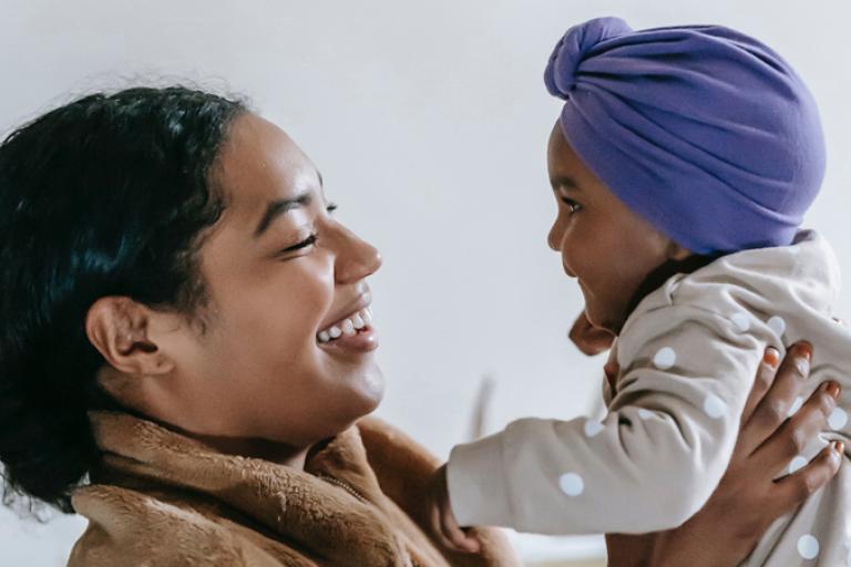 A lady holds a baby facing her, both are smiling at each other