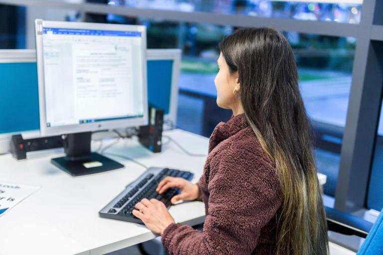 A lady with long hair sitting facing a computer with her hands on the keyboard