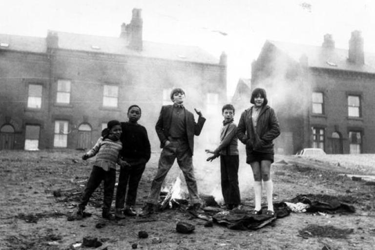 A black and white photo of a group of young people standing near a fire in front of some terraced houses