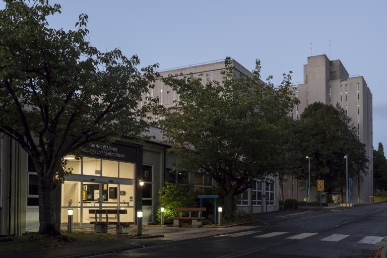 An external view of a building in the dusk. There is a crossing in front of it and trees around it. 