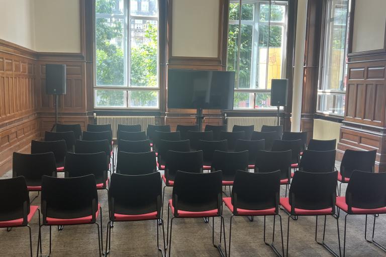 The interior of a wood panelled room with chairs laid out in rows, two big windows at the back and a TV screen