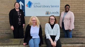 4 member of staff sitting and standing in front of the Leeds School Library Service sign