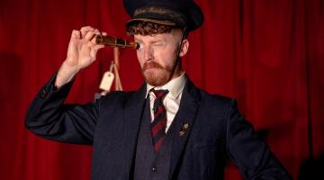 A man wearing a suit and a cap looking through a telescope and standing in front of a red velvet curtain