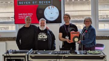 4 people standing behind a turntable smiling at the camera, with one holding a book