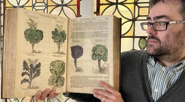 A librarian holding up an old book with illustrations of plants and a stained glass window in the background