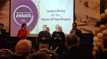 A photo of an awards ceremony - a stage can be seen with 5 people standing in front of a screen. One man is holding the trophy and another is speaking into a microphone