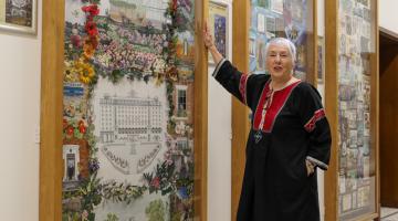 An lady in a black tunic with red trousers points towards a large tapestry panel